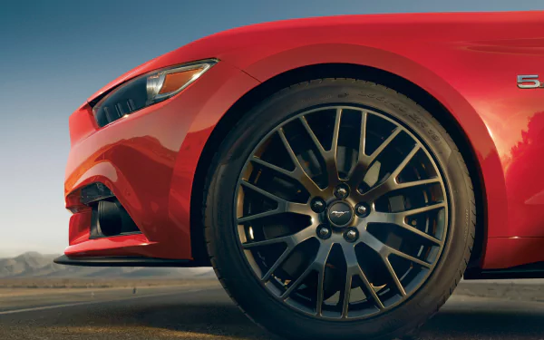 Close-up of a 2015 Ford Mustang GT's wheel, showcasing its sleek design and performance features. The vibrant red car stands against a serene backdrop, highlighting its sporty appeal.