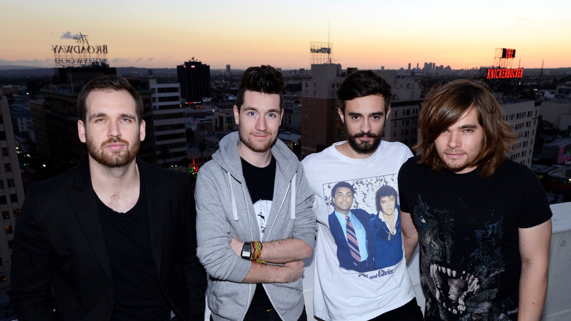 HD desktop wallpaper featuring the English rock band Bastille posing together against a cityscape at sunset, capturing their music and style.