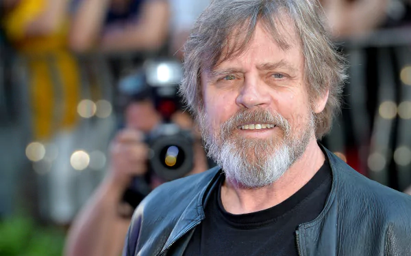 HD desktop wallpaper featuring a smiling Mark Hamill in a black outfit with a camera in the background.