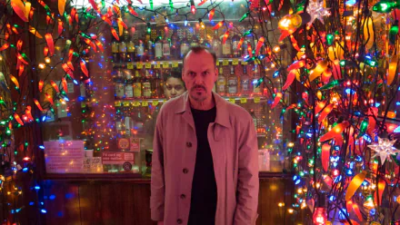 4K Ultra HD PC wallpaper of a brooding actor in a trench coat amid colorful string lights in a cramped bar, evoking the theatrical mood of Birdman.