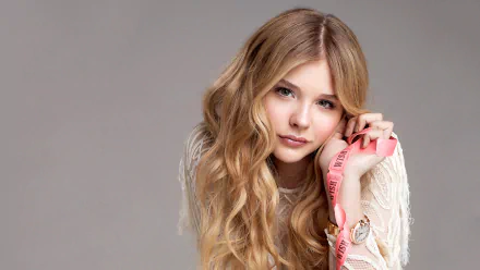 HD desktop wallpaper featuring celebrity Chloë Grace Moretz with long wavy hair, wearing a white top and holding a pink strap against a neutral background.