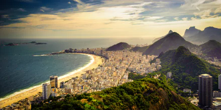 Aerial view of Copacabana beach and Rio de Janeiro cityscape in Brazil, featuring the coastline, urban buildings, and surrounding green hills under a partly cloudy sky.