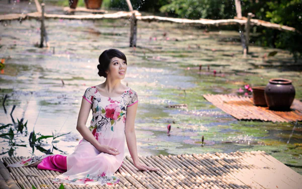 A Vietnamese woman in traditional attire sits gracefully by a serene, water-filled landscape. The HD wallpaper captures her serene expression amid the natural beauty.