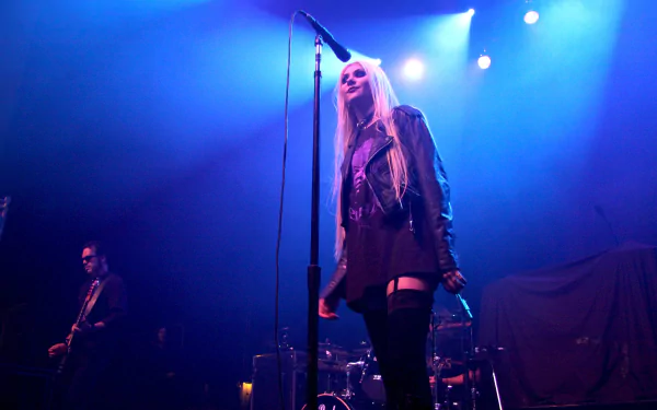 4K Ultra HD PC desktop wallpaper of a female rock singer in a leather jacket at a microphone on a blue-lit stage, with a guitarist and drum kit visible behind her.