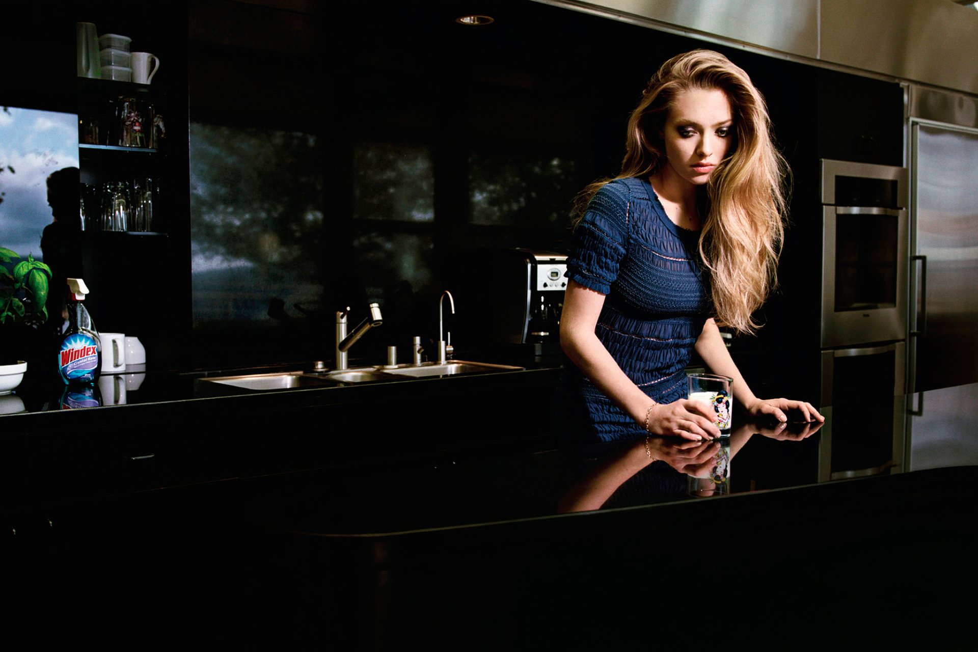 HD PC desktop wallpaper featuring celebrity Amanda Seyfried leaning on a modern kitchen counter with a reflective black surface and dark cabinetry in the background.