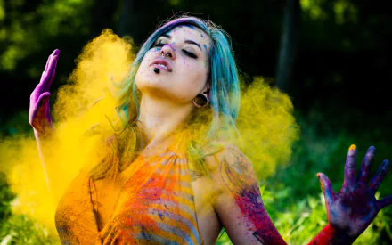 A young person with blue hair and tattoos stands amidst a vibrant display of colors, celebrating Holi, with colorful powders swirling around them, creating a lively and festive mood.