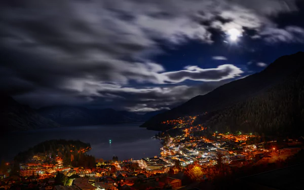 Night view of Queenstown, New Zealand, with glowing town lights beneath a cloudy sky and moon, captured in stunning 4K Ultra HD resolution.