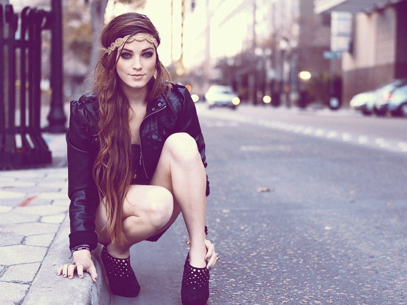 HD PC desktop wallpaper of a woman model crouching on an urban street, long hair, leather jacket, blurred cars and buildings in the background.