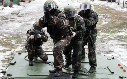 Military HD PC desktop wallpaper: four camouflaged soldiers in full combat gear and helmets on an armored vehicle, aiming rifles over snowy ground.