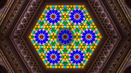 A vibrant stained glass pattern featuring blue and yellow floral designs, set in a hexagonal frame, showcasing intricate architectural details of a roof at Vijecnica.