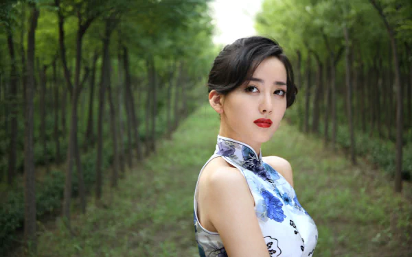 A stunning Chinese woman, Hú Yǐng Yí, poses gracefully amidst a lush green forest, showcasing elegance in her floral-patterned dress. This vibrant image serves as a captivating 4K Ultra HD background.