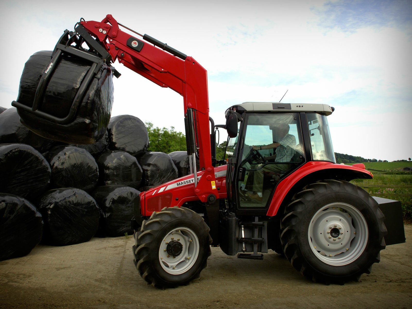 A Massey Ferguson tractor lifts a large black bale in front of a stack, set against a scenic backdrop. This HD image serves as an impressive desktop wallpaper.