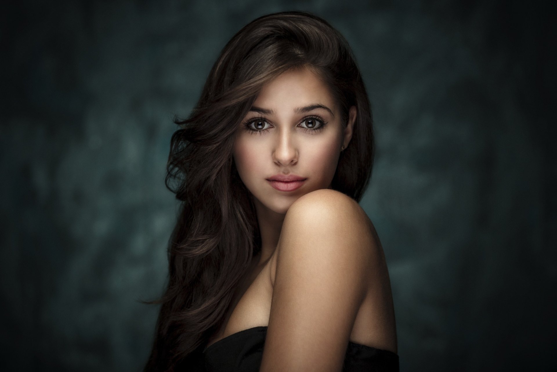 A striking HD wallpaper featuring a brunette woman with soft, wavy hair, gazing confidently at the viewer against a moody background. A captivating desktop background choice.