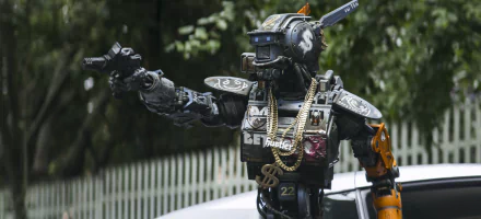 HD desktop wallpaper featuring Chappie, the robot character with gold chains, gesturing in front of a vehicle.