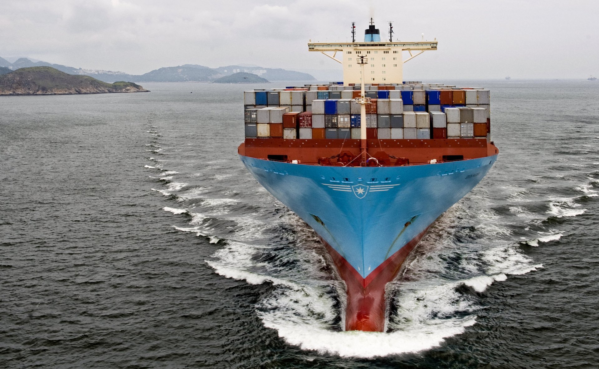 HD desktop wallpaper showing a large container ship navigating open waters with stacked cargo containers against a cloudy sky and distant hills.