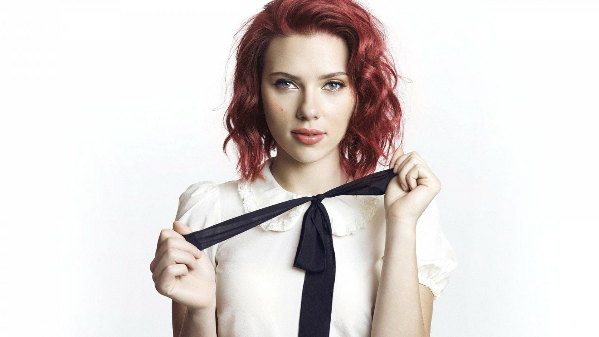 Scarlett Johansson poses with a playful expression, holding the tie of her blouse. This HD wallpaper captures her vibrant personality against a clean white backdrop.