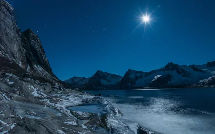 HD desktop wallpaper of a serene night landscape featuring a bright starry sky, moonlit rocky shore, and distant mountains reflecting on calm waters.