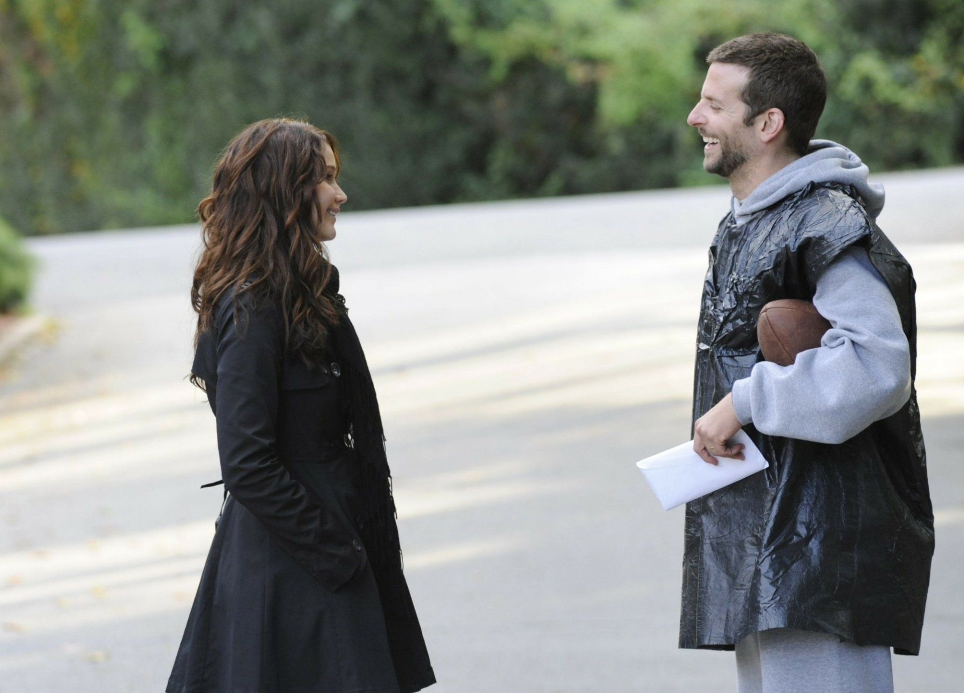 HD desktop wallpaper featuring Bradley Cooper and Jennifer Lawrence in a scene from the movie Silver Linings Playbook, smiling and conversing outdoors.