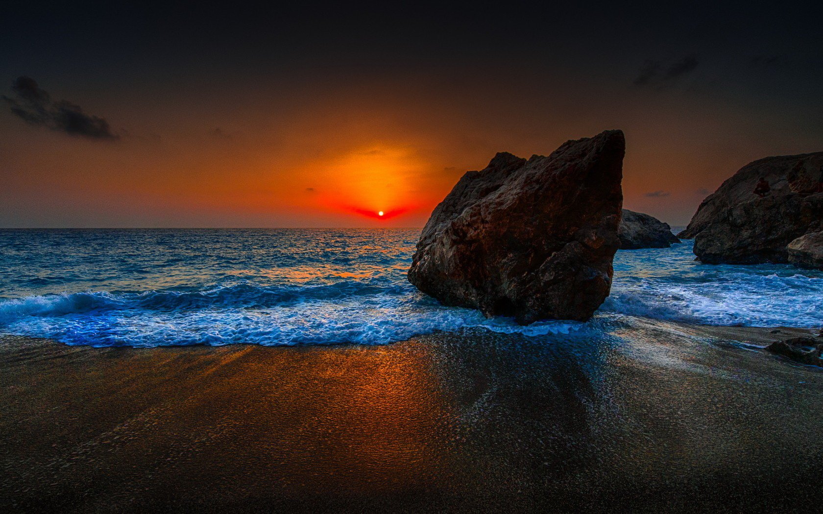 HD PC desktop wallpaper/background: nature sunset over a rocky shoreline — glowing orange sun dipping below the horizon, blue waves washing a dark sandy beach.
