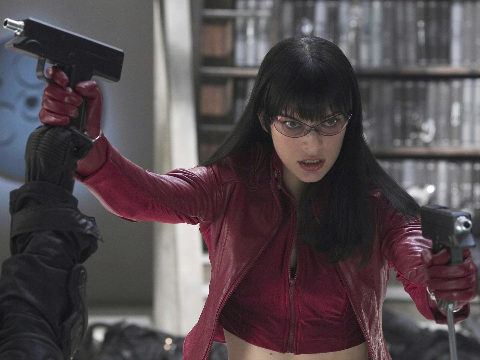 HD PC desktop wallpaper from the movie Ultraviolet: dark-haired female character in a red leather jacket and glasses wielding twin pistols in a library scene.