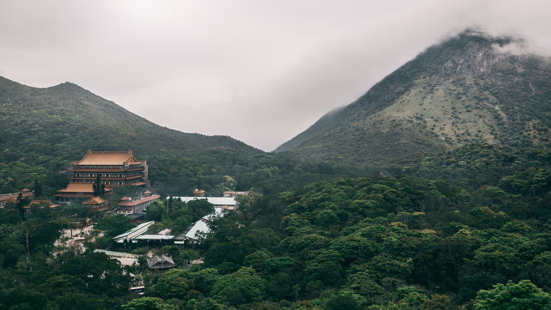 Fog blankets the mountainous landscape of Lantau Island, Hong Kong, featuring the serene Po Lin Monastery nestled among lush greenery.