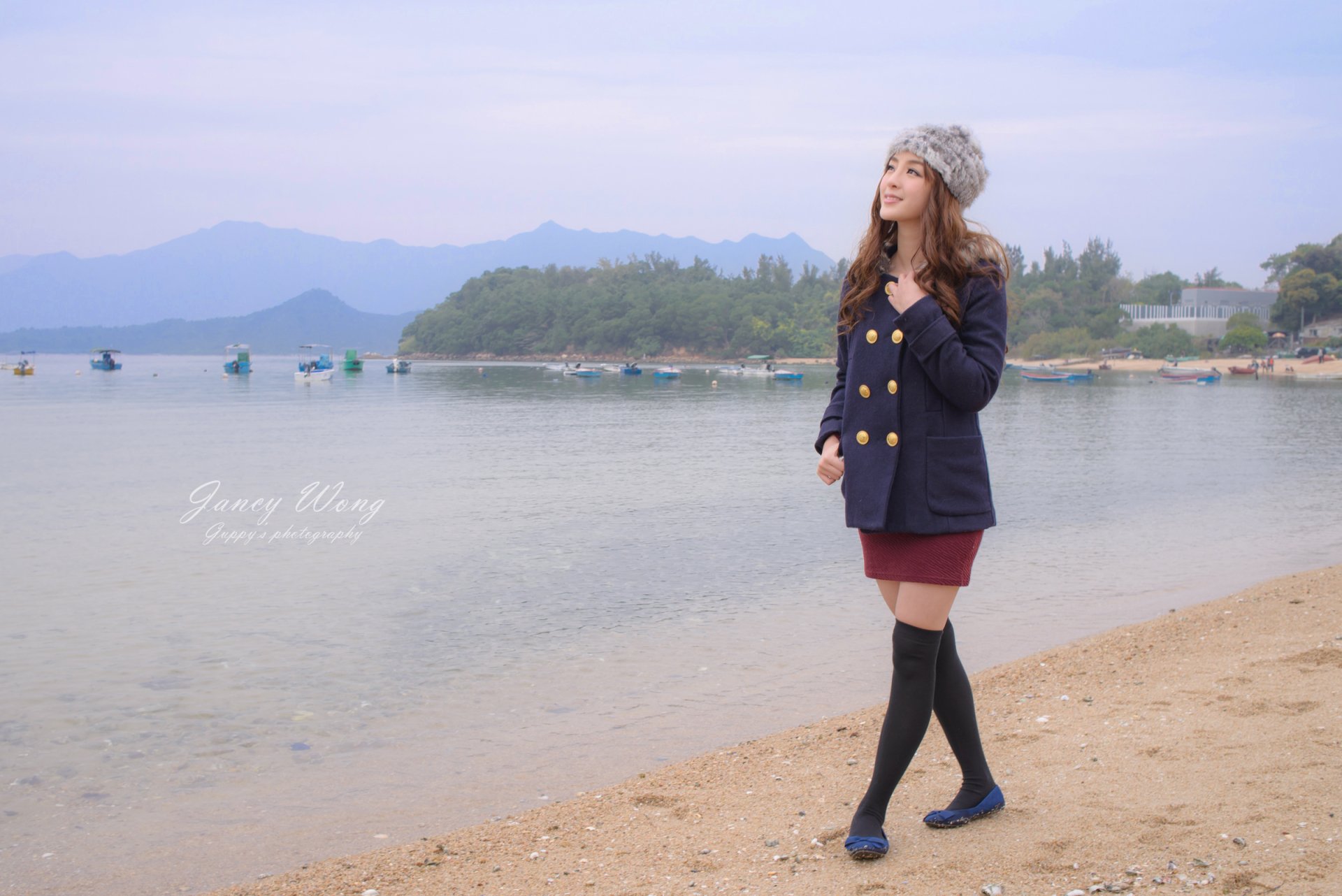 HD desktop wallpaper featuring an Asian woman model, Jancy Wong, smiling by the beach wearing a cap, navy coat, and boots, with mountains and boats in the background.