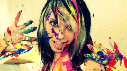 Colorful artistic HD PC desktop wallpaper and background of a young woman with paint-splattered face and hands, tongue out, vibrant streaks of color across hair and skin.
