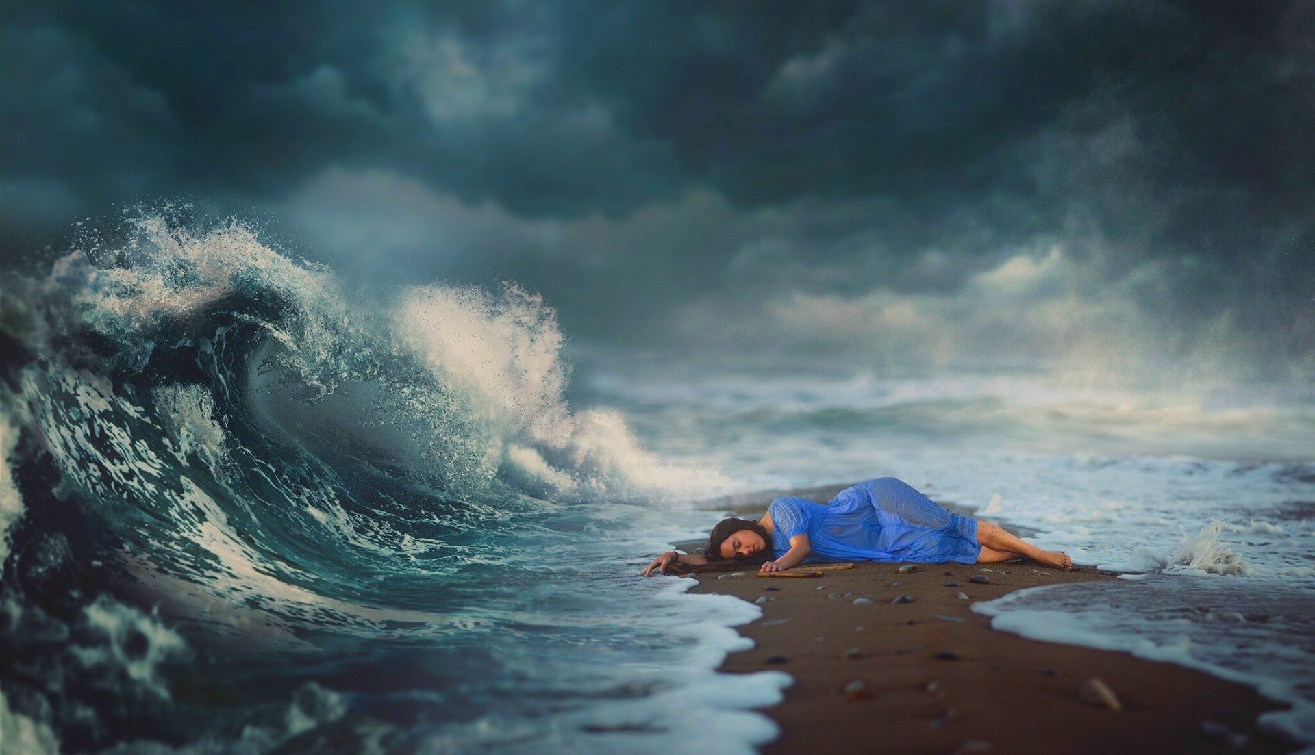 A woman in a blue dress lies on a beach as waves crash nearby under a stormy sky. HD desktop wallpaper and background.