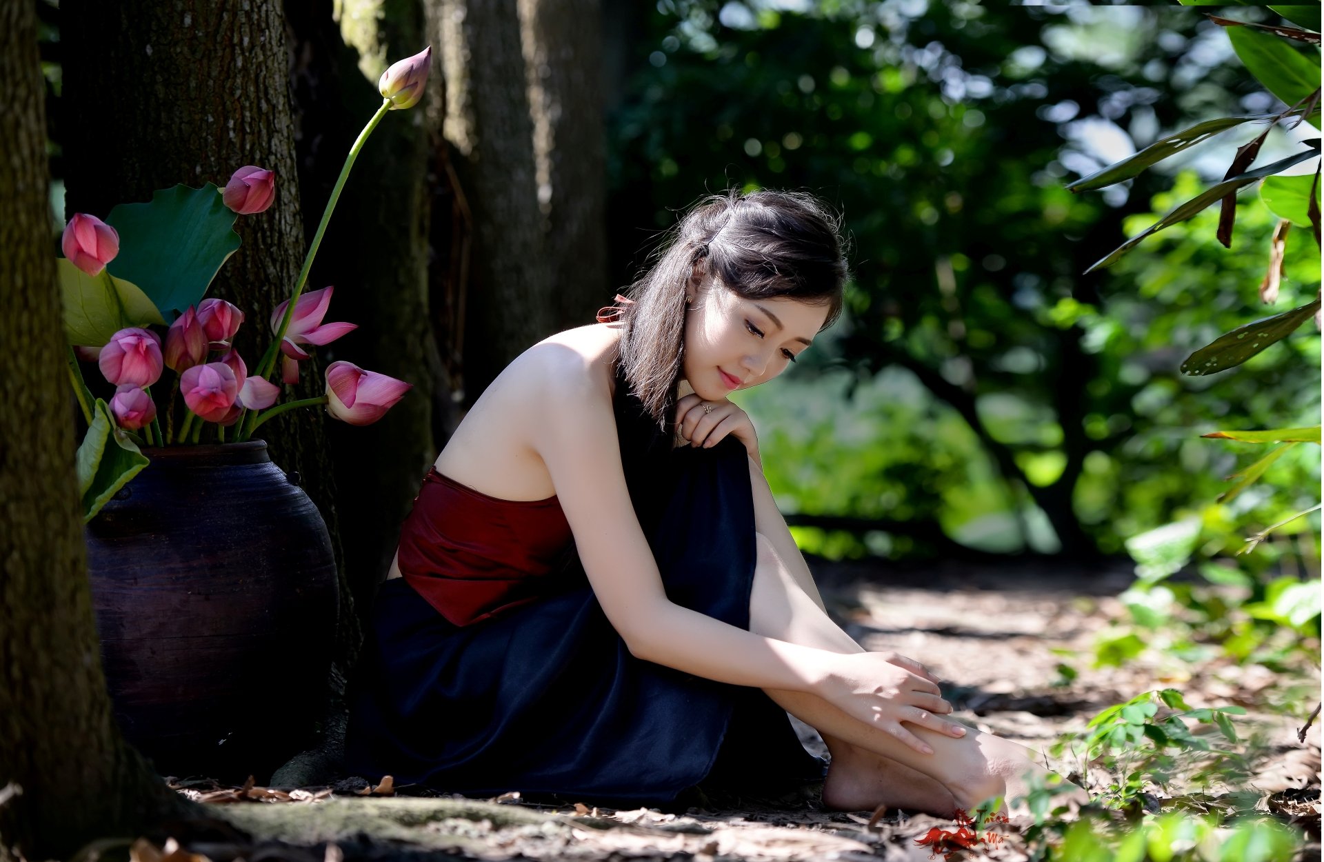 A serene image of an Asian woman sitting on the ground in a forested area near a vase of lotus flowers. The background features bokeh, adding a dreamy effect. HD desktop wallpaper and background.