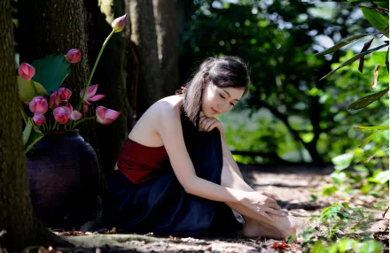 A serene image of an Asian woman sitting on the ground in a forested area near a vase of lotus flowers. The background features bokeh, adding a dreamy effect. HD desktop wallpaper and background.