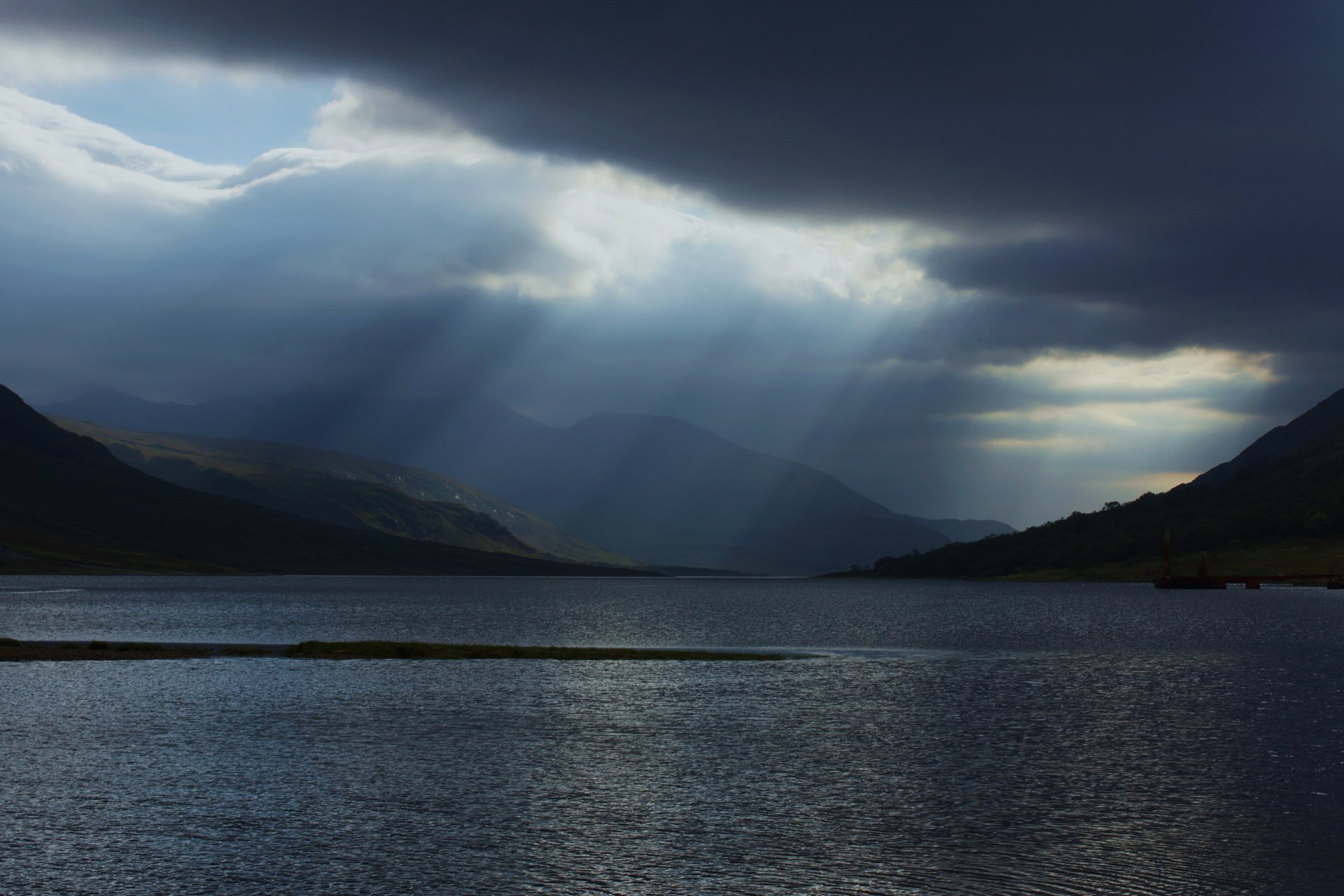 Sunbeams break through dark clouds over Loch Etive, Scotland, casting light on the serene lake surrounded by misty hills in this 4K Ultra HD nature wallpaper.
