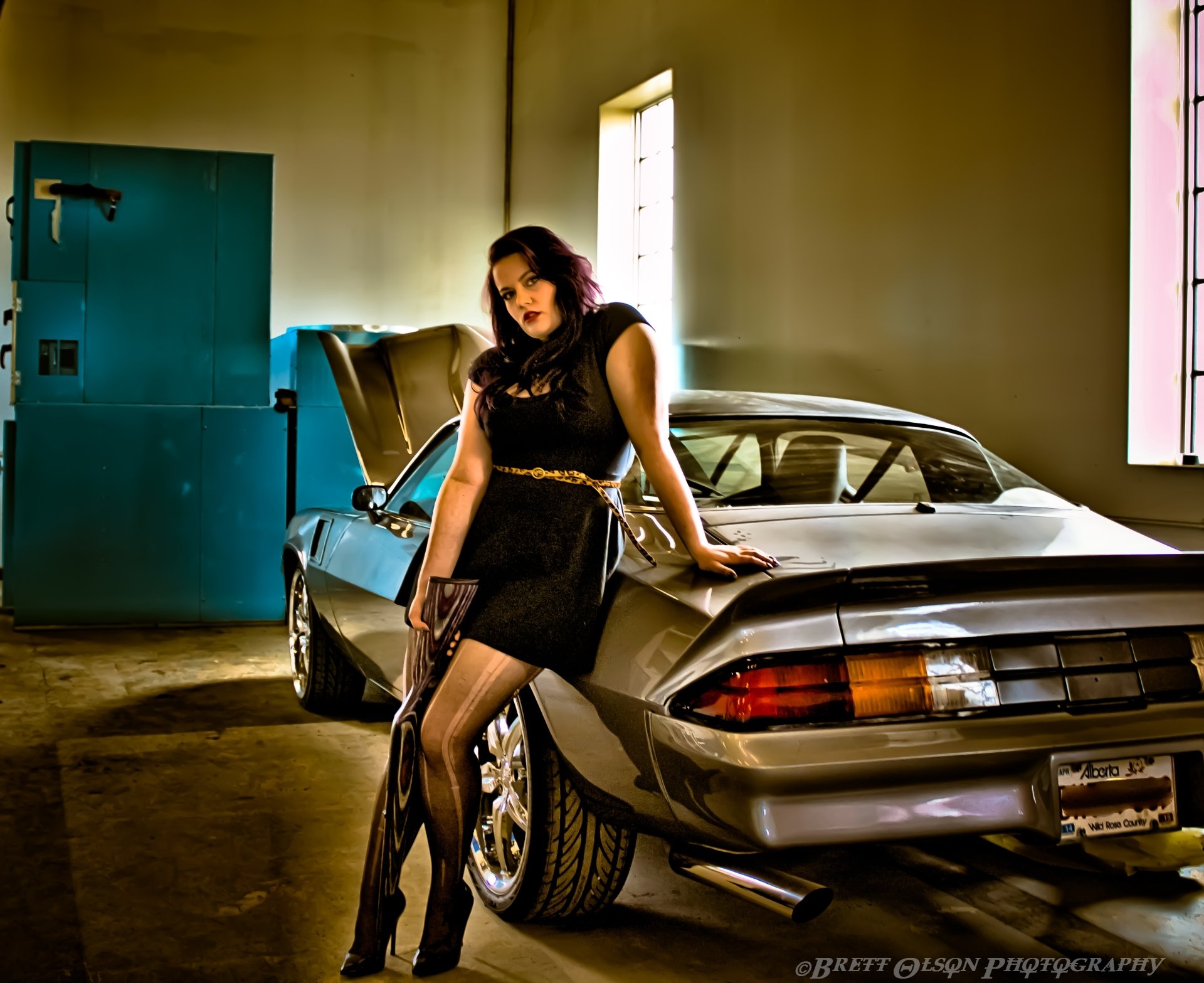 HD desktop wallpaper featuring a woman in a black dress leaning against a silver Chevrolet Camaro, holding a gun in an indoor garage setting.