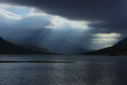 Sunbeams break through dark clouds over Loch Etive, Scotland, casting light on the serene lake surrounded by misty hills in this 4K Ultra HD nature wallpaper.
