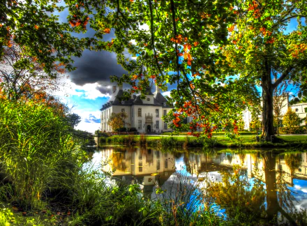 A serene view of Castle Gartrop in North Rhine-Westphalia, Germany, reflecting on a tranquil lake surrounded by vibrant trees and lush greenery.