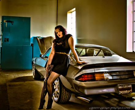 HD desktop wallpaper featuring a woman in a black dress leaning against a silver Chevrolet Camaro, holding a gun in an indoor garage setting.