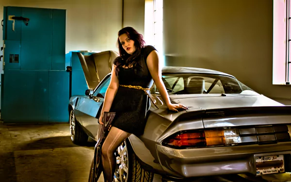 HD desktop wallpaper featuring a woman in a black dress leaning against a silver Chevrolet Camaro, holding a gun in an indoor garage setting.