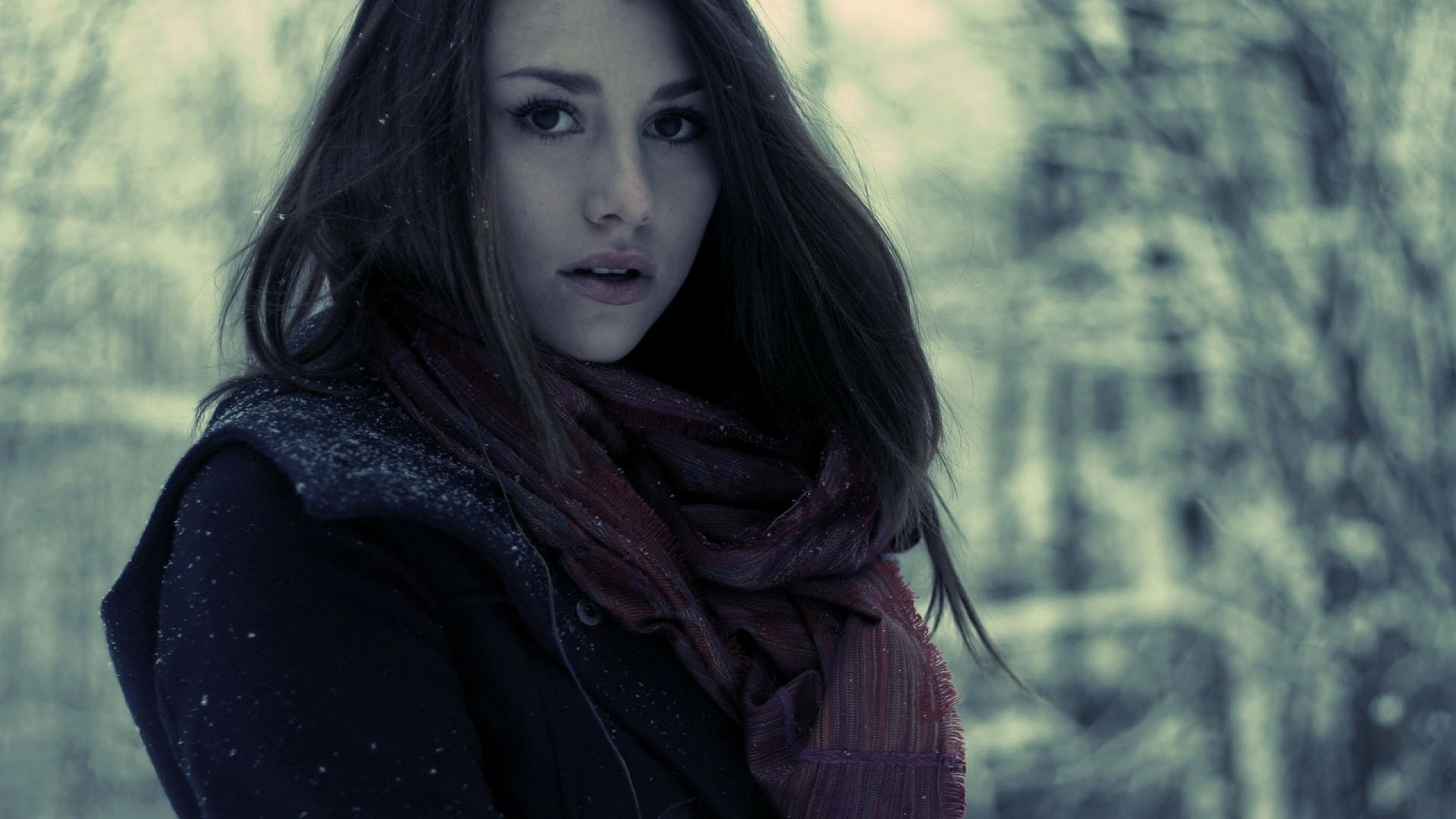 HD PC desktop wallpaper featuring a beautiful brunette woman with long hair, wearing a dark coat and scarf, set against a blurred winter forest background.