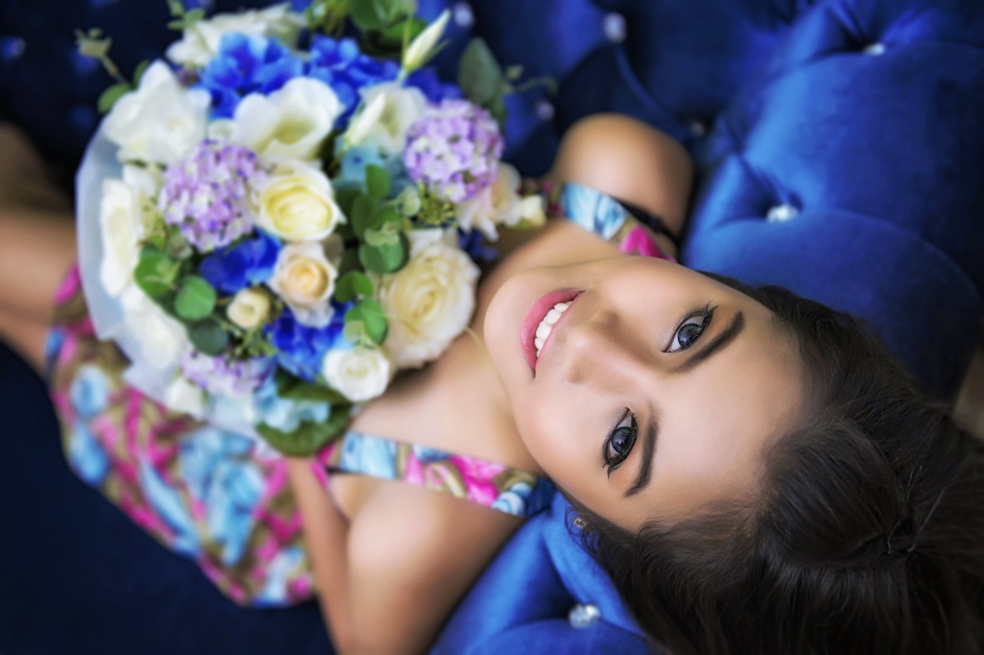 A Thai woman smiles brightly, holding a bouquet of colorful flowers, seated against a plush blue background. This vibrant image captures beauty and joy in stunning 4K Ultra HD.
