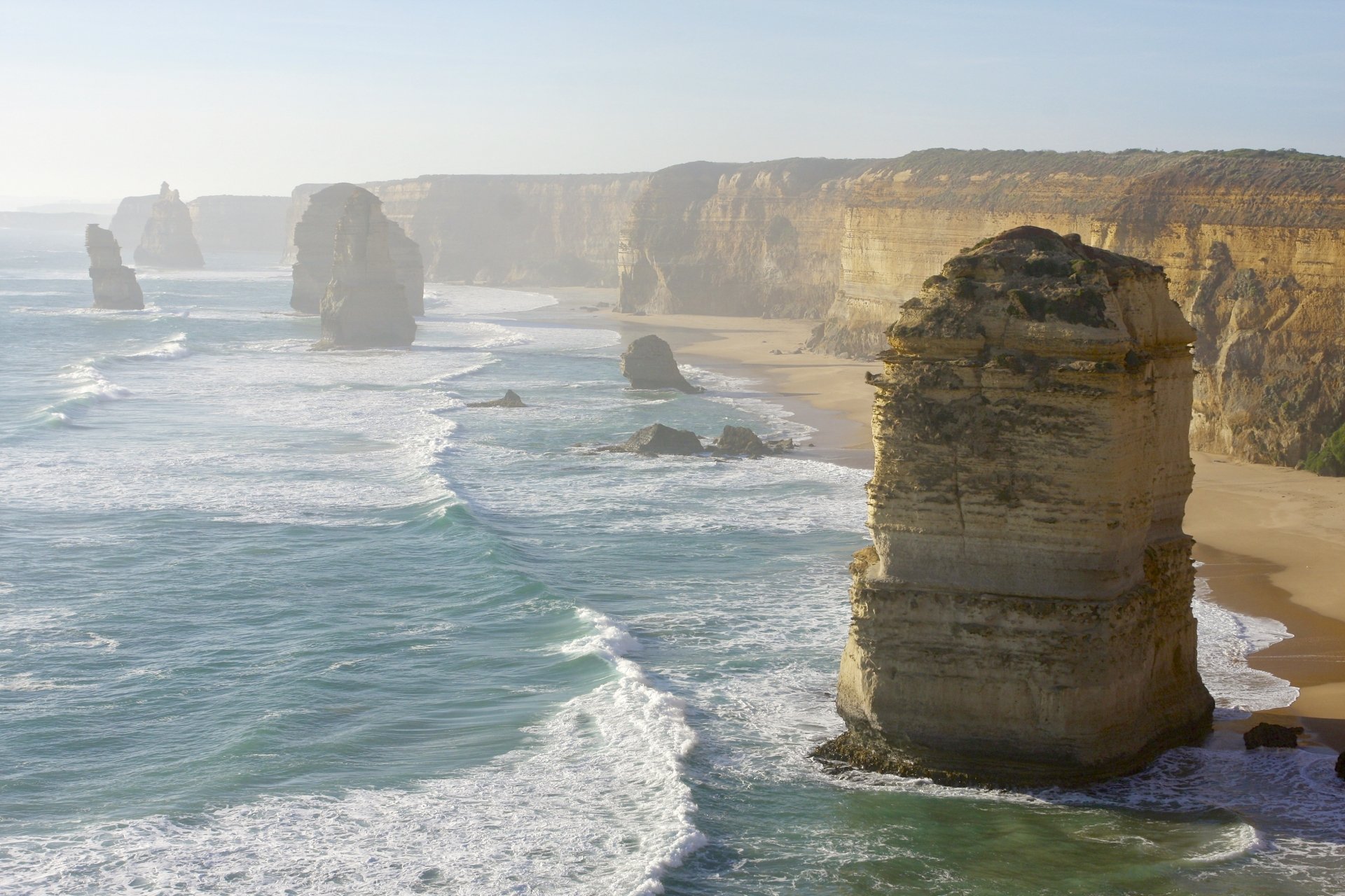 A 4K Ultra HD view of the Twelve Apostles limestone stacks along Victoria, Australia's coastline with waves crashing against the rugged cliffs and sandy shore.