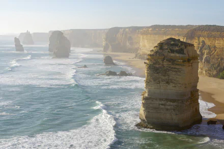 A 4K Ultra HD view of the Twelve Apostles limestone stacks along Victoria, Australia's coastline with waves crashing against the rugged cliffs and sandy shore.