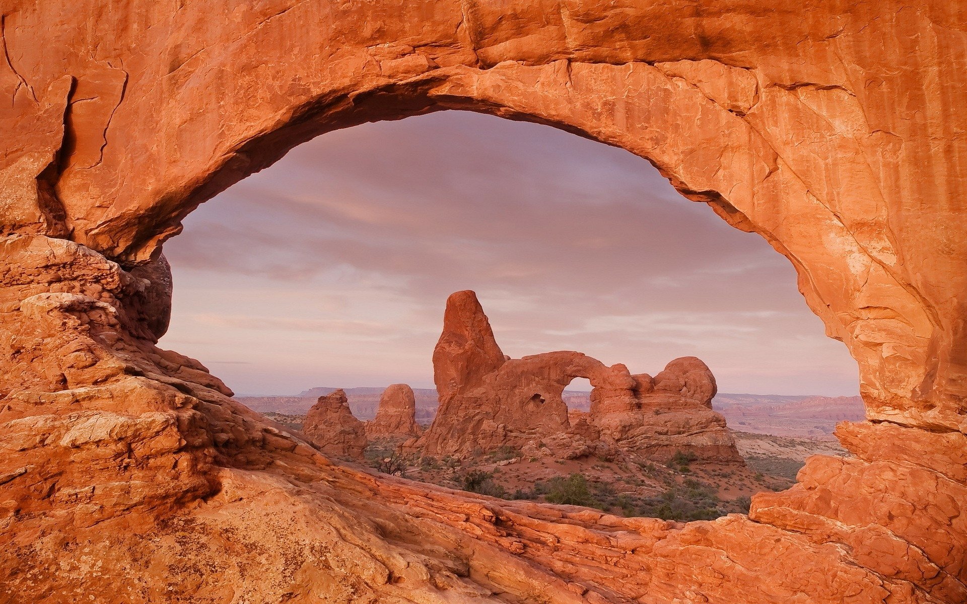 HD desktop wallpaper showcasing a natural desert landscape with a prominent rock arch framing distant sandstone formations under a soft, pastel sky.