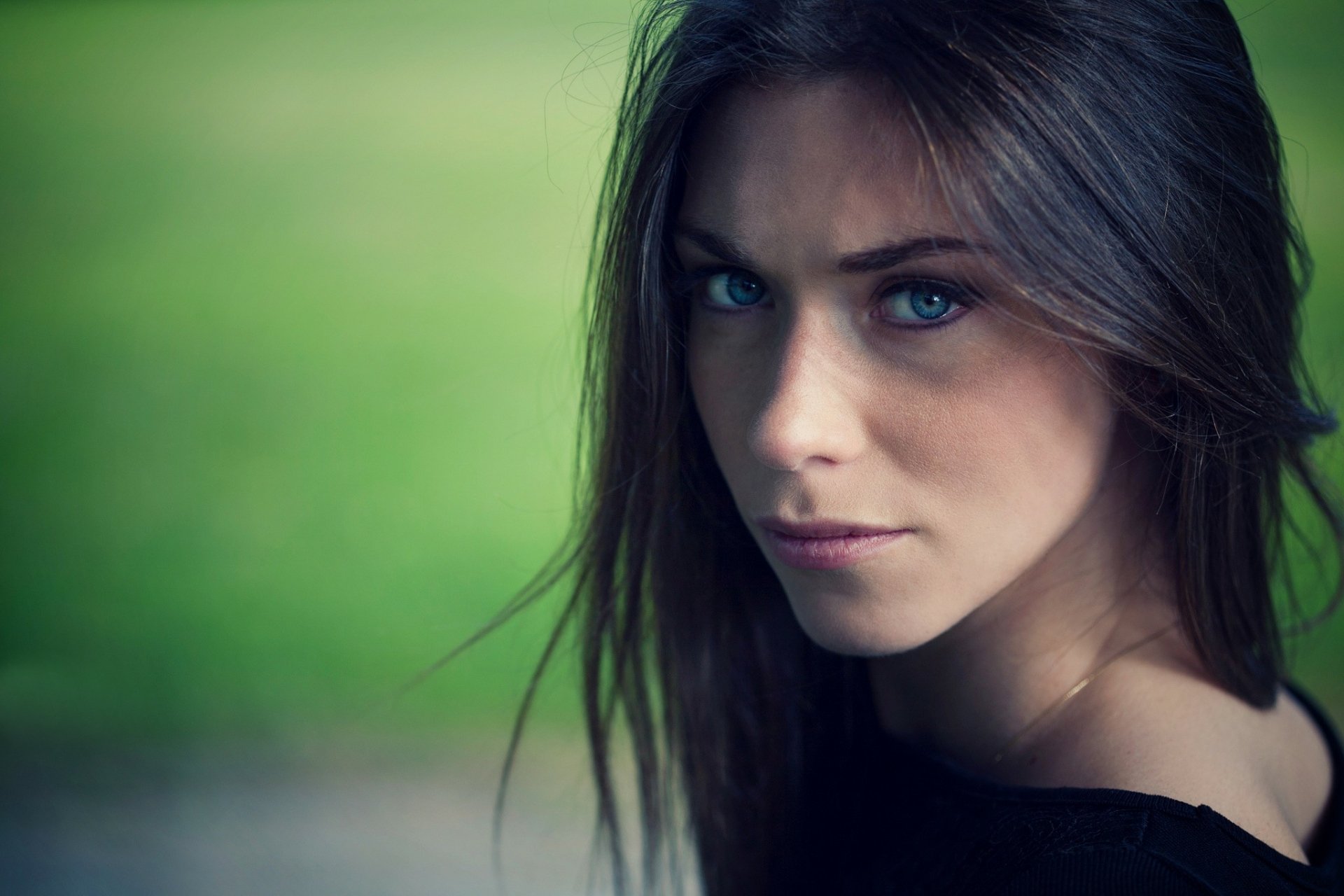A close-up portrait of a brunette woman with striking blue eyes, set against a softly blurred green background, creating an engaging HD desktop wallpaper.
