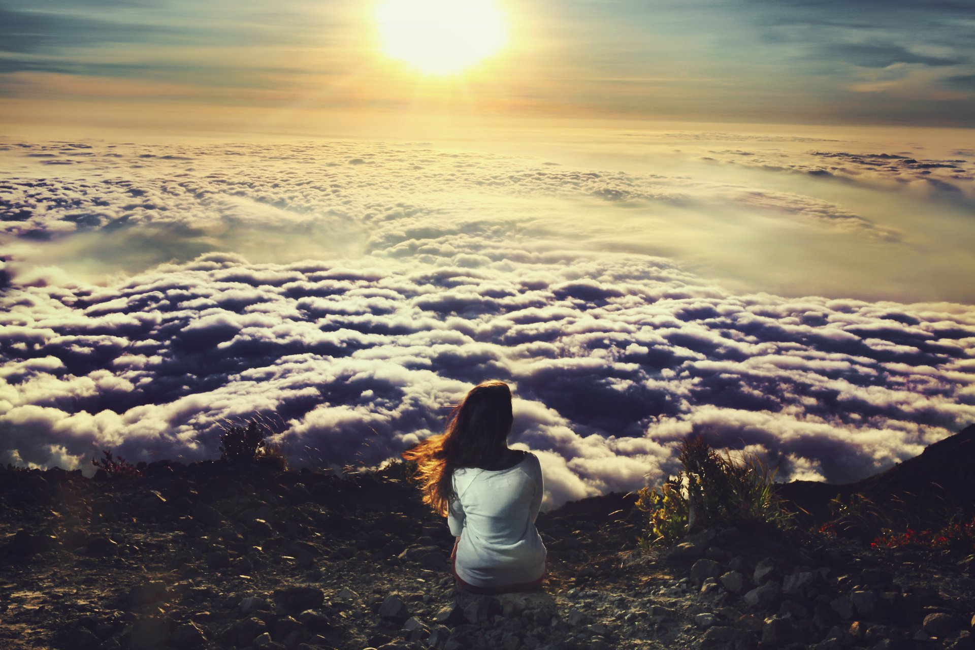 A brunette woman sits on rocky terrain, gazing at a breathtaking landscape of clouds bathed in golden sunlight, creating a serene and captivating atmosphere.