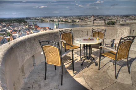 A panoramic view from Halászbástya in Budapest, showcasing the Danube River and the Hungarian Parliament Building, with a terrace featuring a table and chairs.