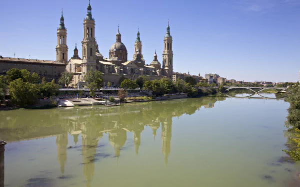 A stunning 4K Ultra HD view of the Cathedral-Basilica of Our Lady of the Pillar by the Ebro River in Zaragoza, Spain, showcasing its majestic towers and serene river reflection.