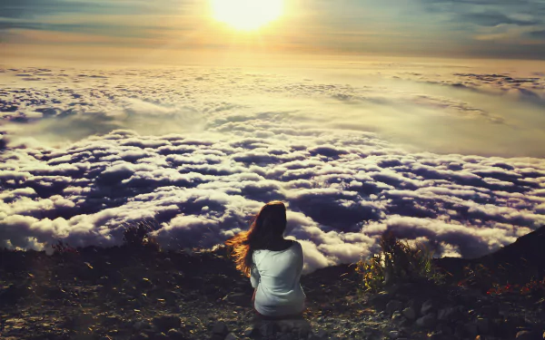 A brunette woman sits on rocky terrain, gazing at a breathtaking landscape of clouds bathed in golden sunlight, creating a serene and captivating atmosphere.