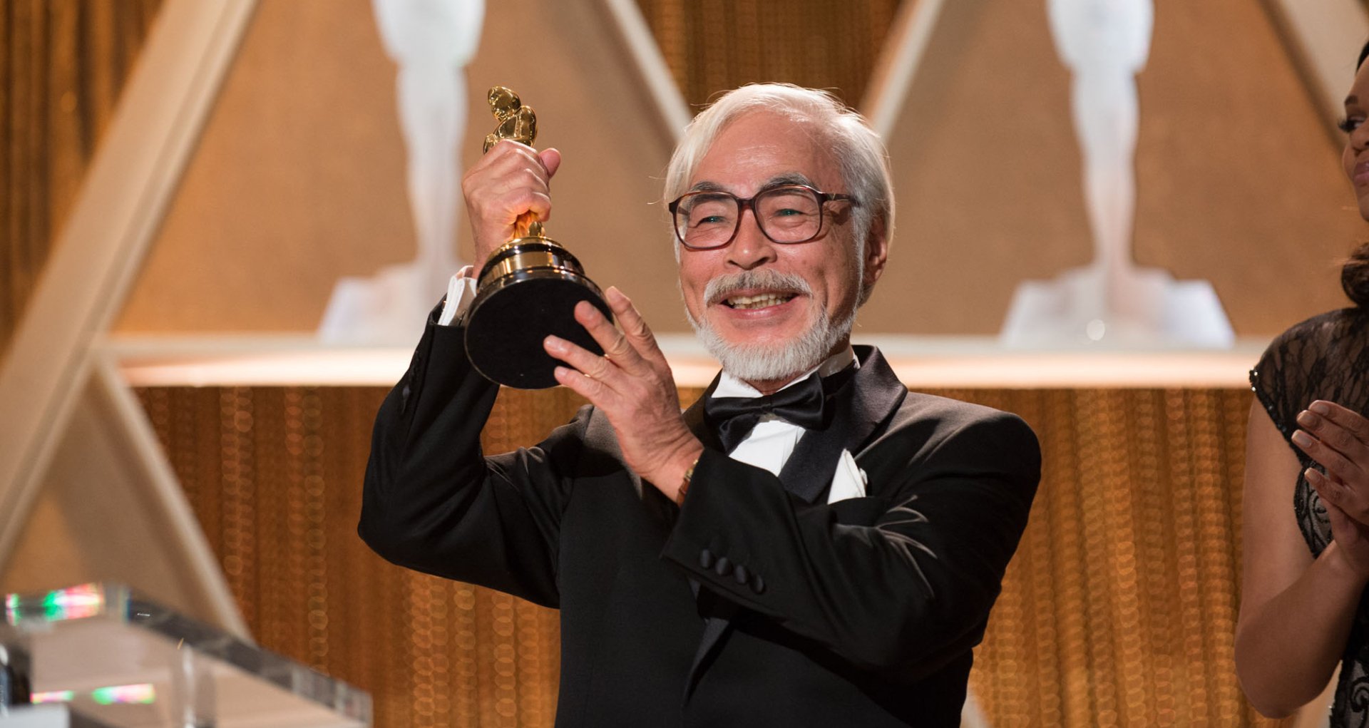 Hayao Miyazaki holding an Oscar, in a tuxedo, with a joyful expression; a high-definition desktop wallpaper.
