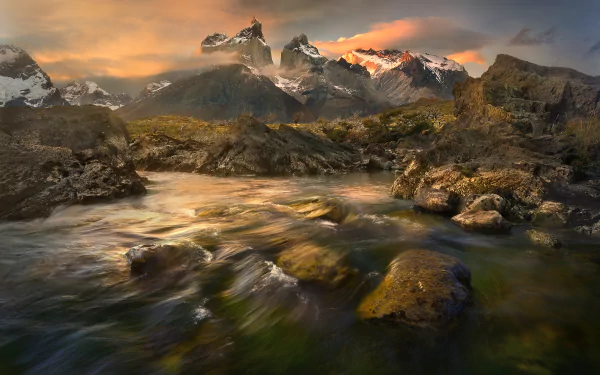 A serene stream flows over rocks in Torres del Paine National Park, Chile, with majestic mountains and a vibrant sunset in the background, showcasing the stunning beauty of Patagonia's nature.