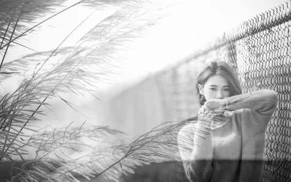 Black & white photo of a Taiwanese model standing by a fence, with tall grass in the background. The model, identified as Lín Yǔ, covers her mouth with her sweater, creating a serene and artistic atmosphere.