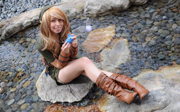 A woman in cosplay poses on a rock by a stream, dressed in a green outfit with brown boots and holding a small item. The vibrant scene makes for an eye-catching HD desktop wallpaper.